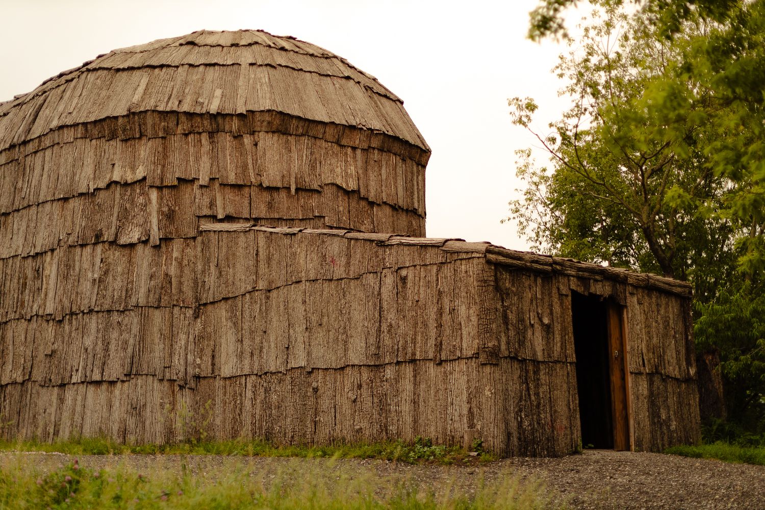 Traditional dome-shaped indigenous longhouse with layered bark exterior beside a rectangular structure in a natural setting.