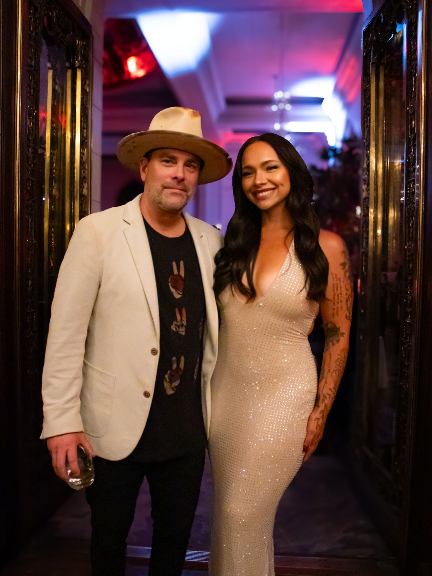 Man in white blazer and tan hat posing with tattooed woman in silver sequin gown at an elegant event.