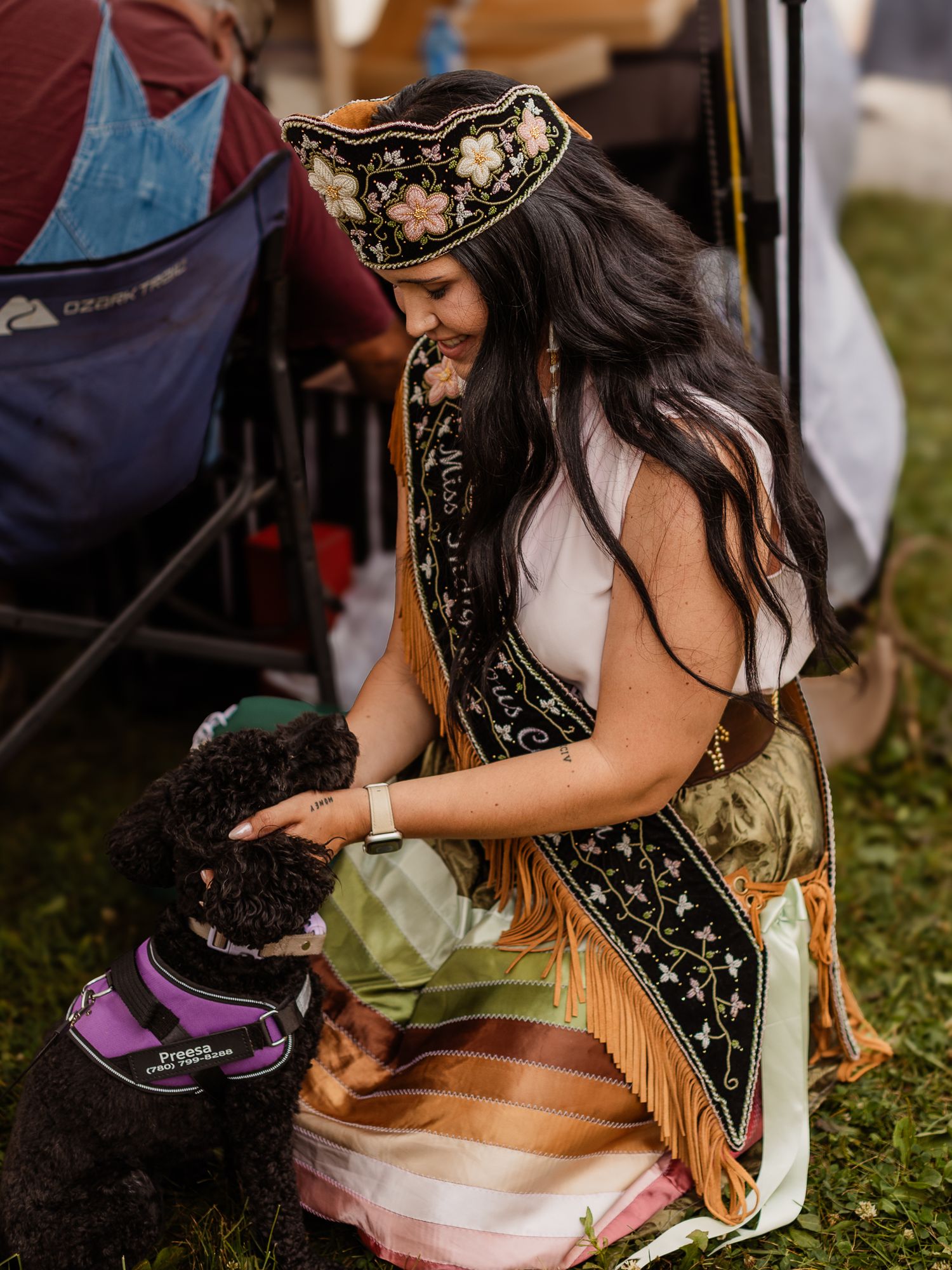 Miss Indigenous Canada titleholder in traditional regalia petting a black poodle service dog.