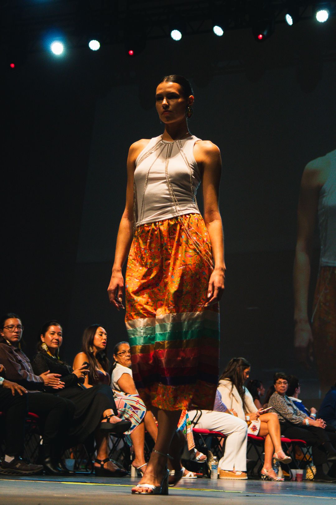 Model walking runway in white halter top and vibrant multicolor midi skirt at a fashion show.