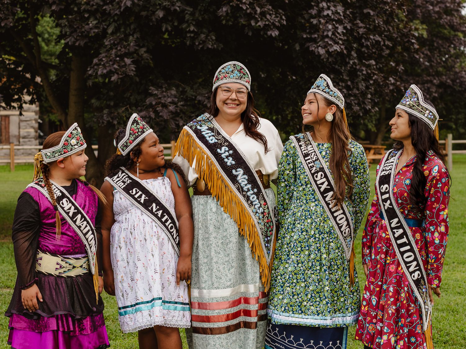 Five women in traditional Indigenous regalia and Six Nations sashes smile together outdoors at a cultural pageant event.