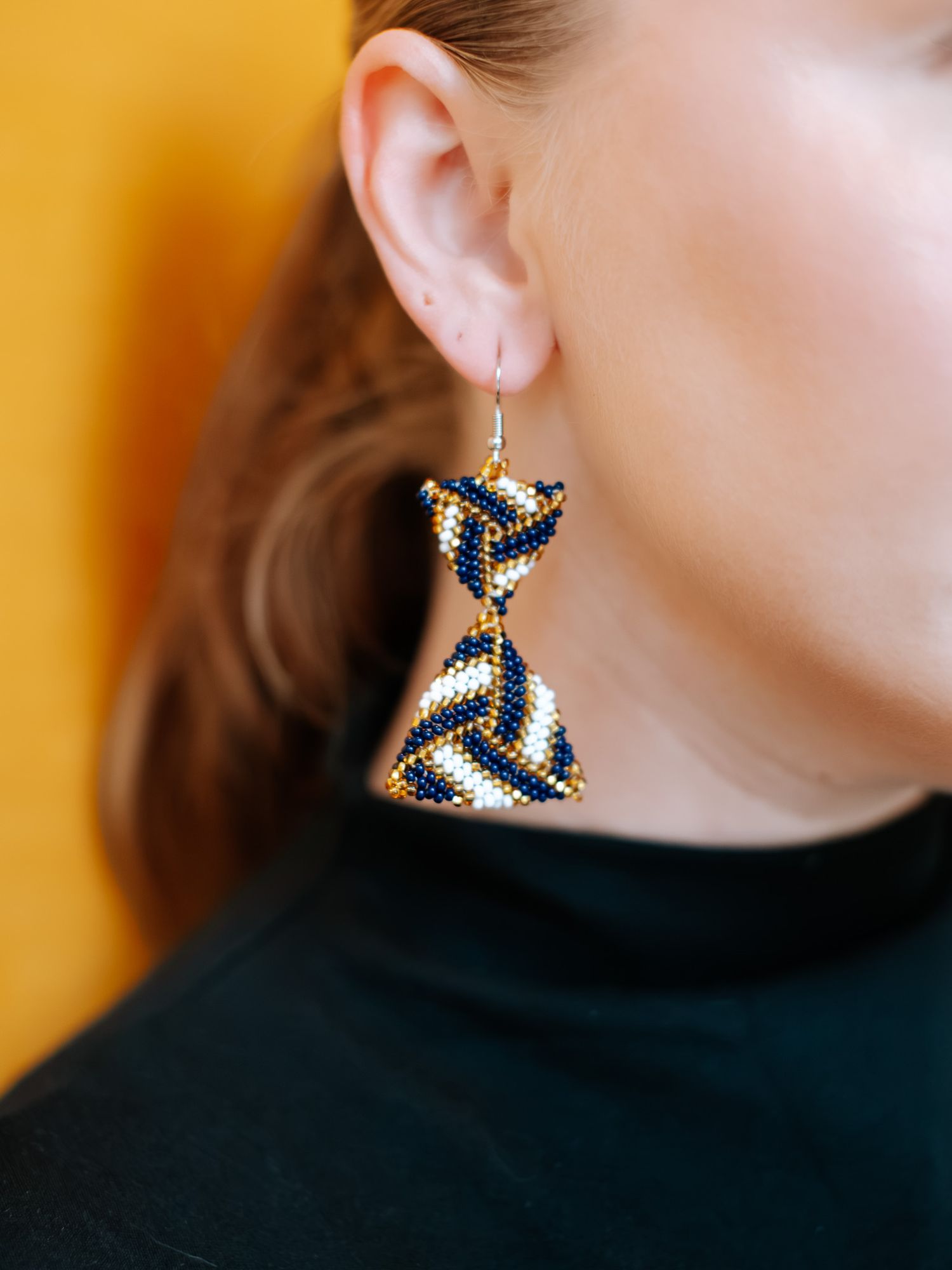 Woman wearing handcrafted beaded earrings in navy blue, white, and gold twisted geometric pattern against yellow background.