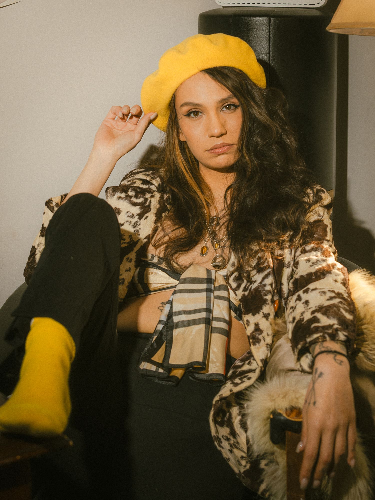 Young woman in yellow beret, cow-print coat, and plaid crop top seated in a stylish indoor setting.