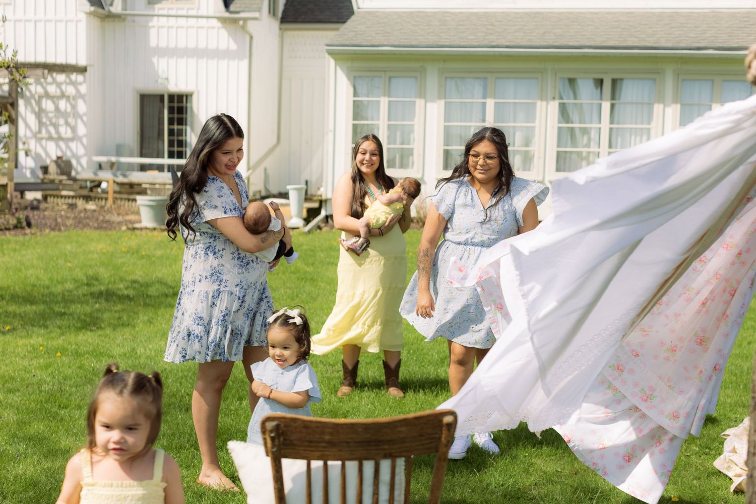 Three women holding babies laugh outdoors near a clothesline, while a toddler plays nearby in a sunny backyard.