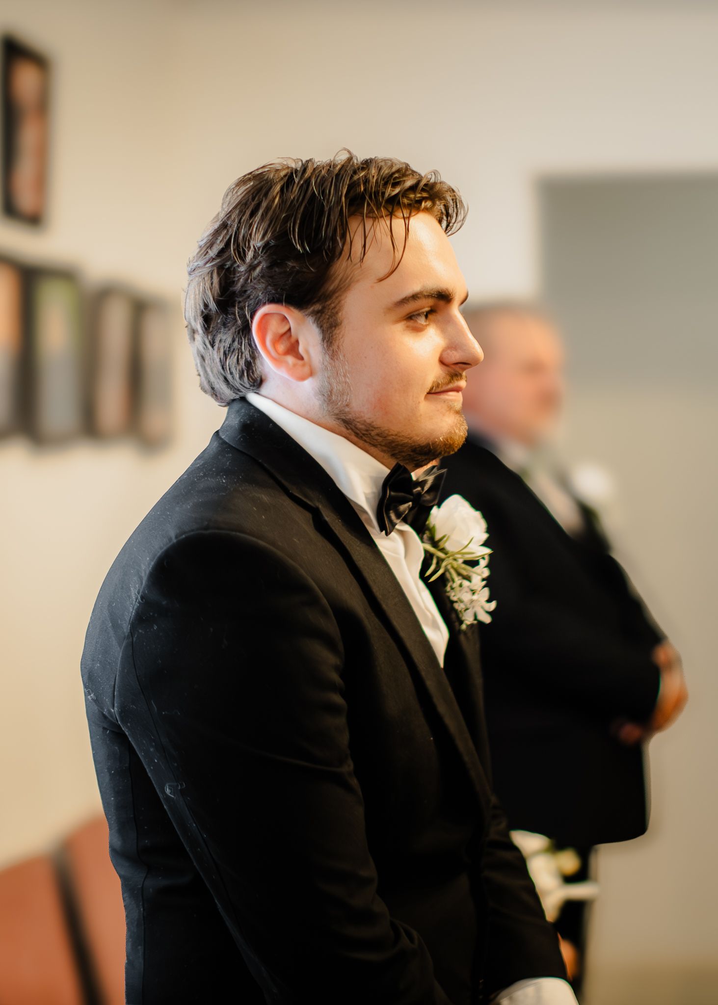 Young groom in black tuxedo with white boutonniere watches bride walk down aisle at wedding ceremony.