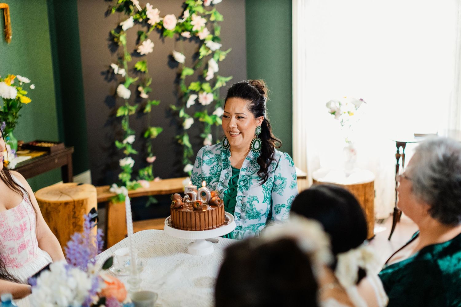 Woman smiling at birthday cake with lit candles at a floral-decorated indoor gathering.