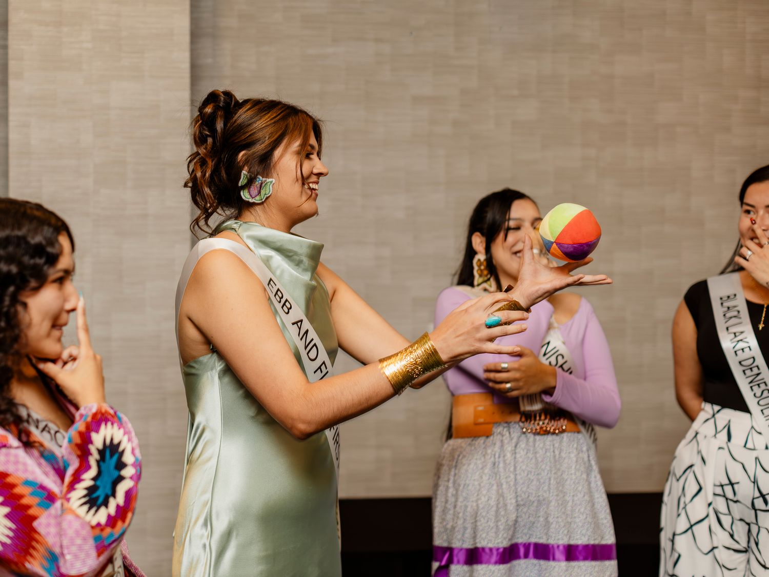 Women in sashes and colorful outfits laugh together at a pageant event, one holding a rainbow ball.