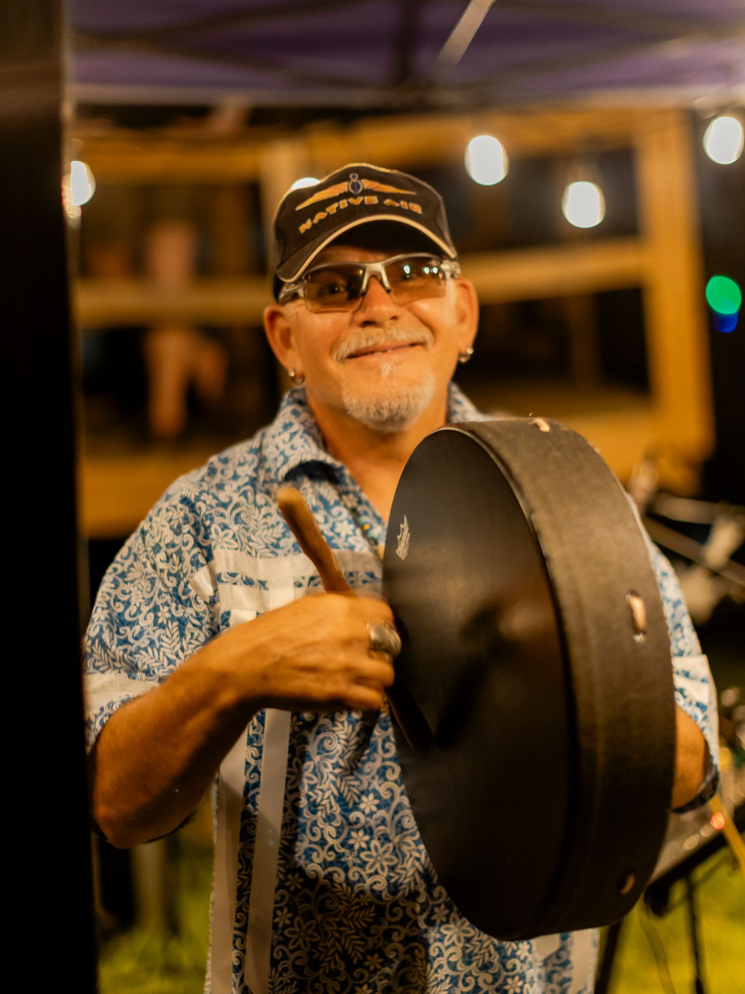 Smiling musician holding a large gong with drumstick at an outdoor evening music event.