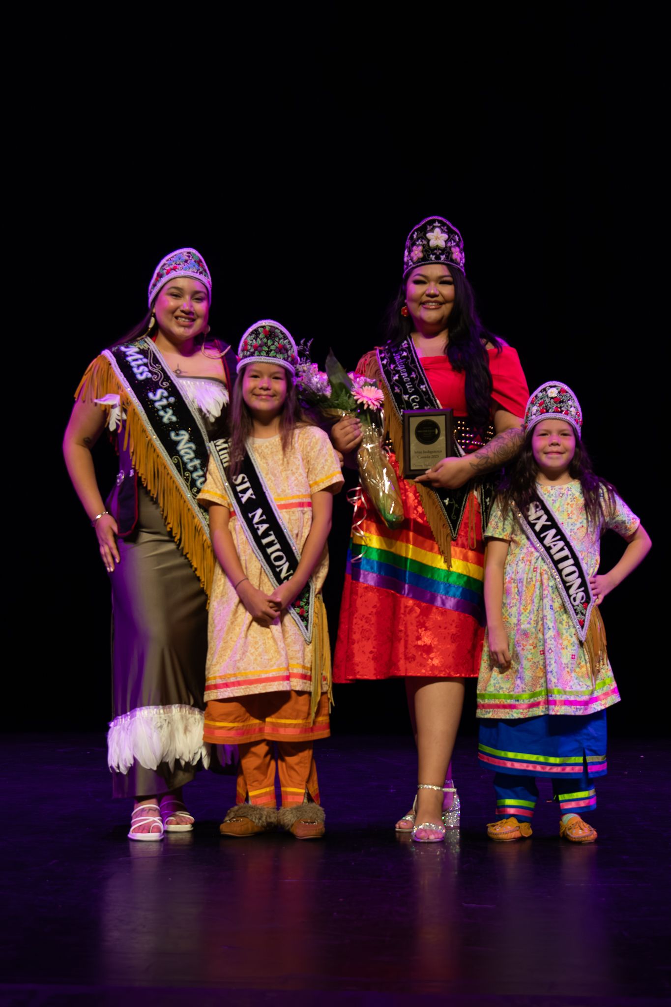 Traditional pageant winners wearing colorful regalia and sashes on stage.