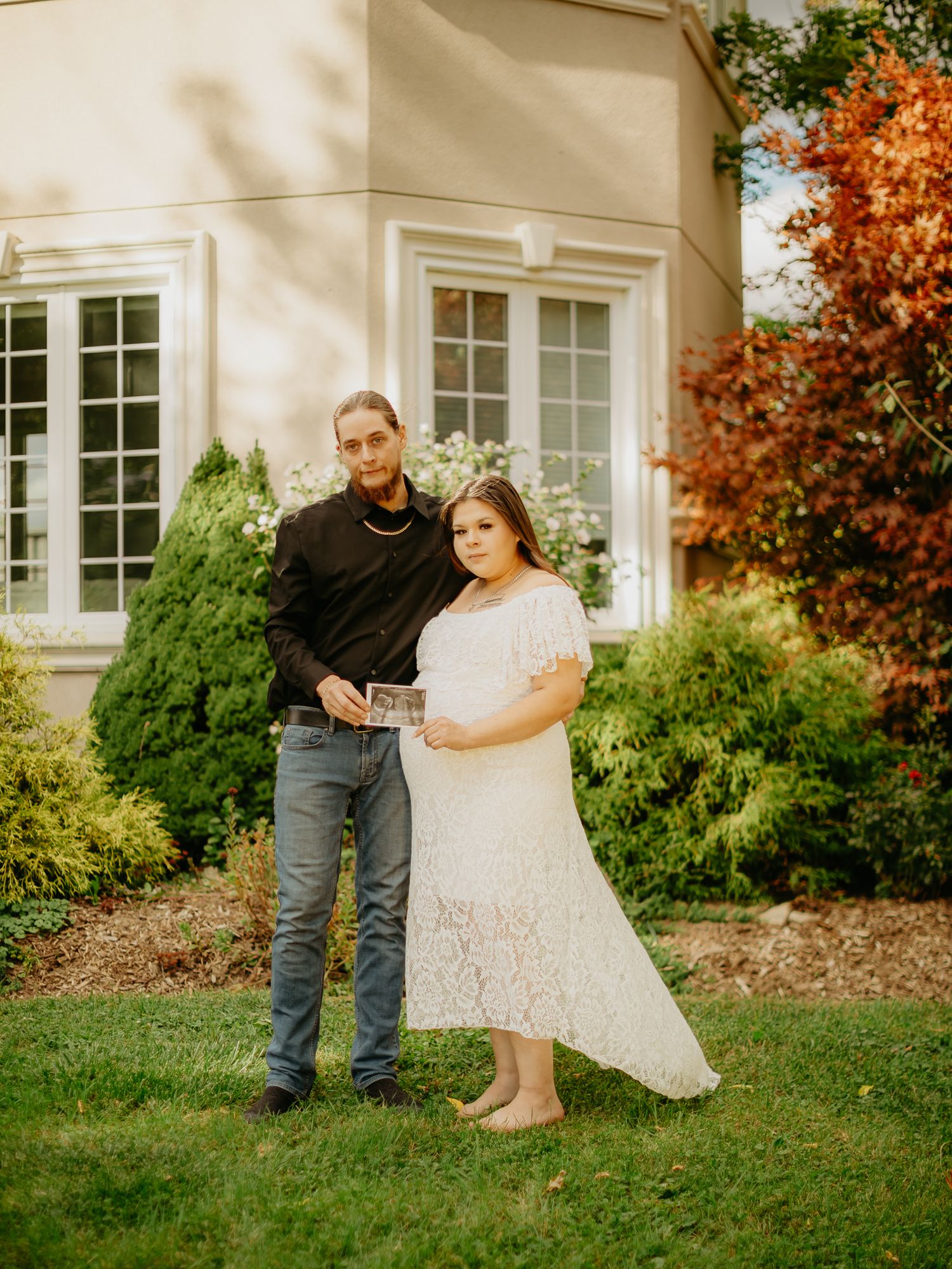 A couple poses together on a lawn in front of a house with white trim and autumn trees.