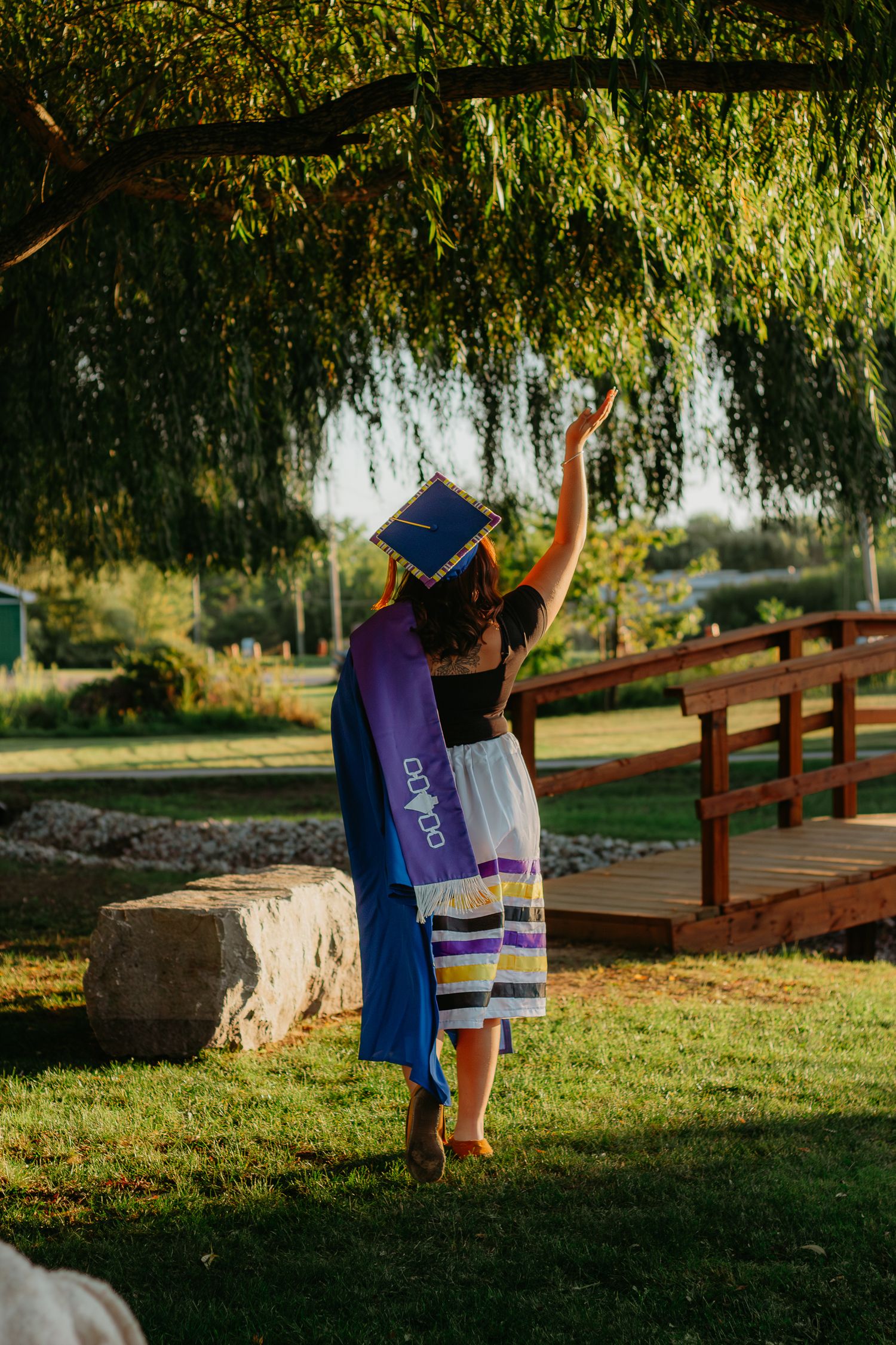 A graduate celebrates near a wooden bridge and pond during a golden sunset.