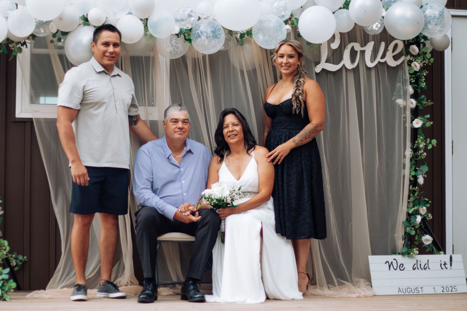 Happy wedding couple poses with two guests in front of Love balloon arch backdrop.