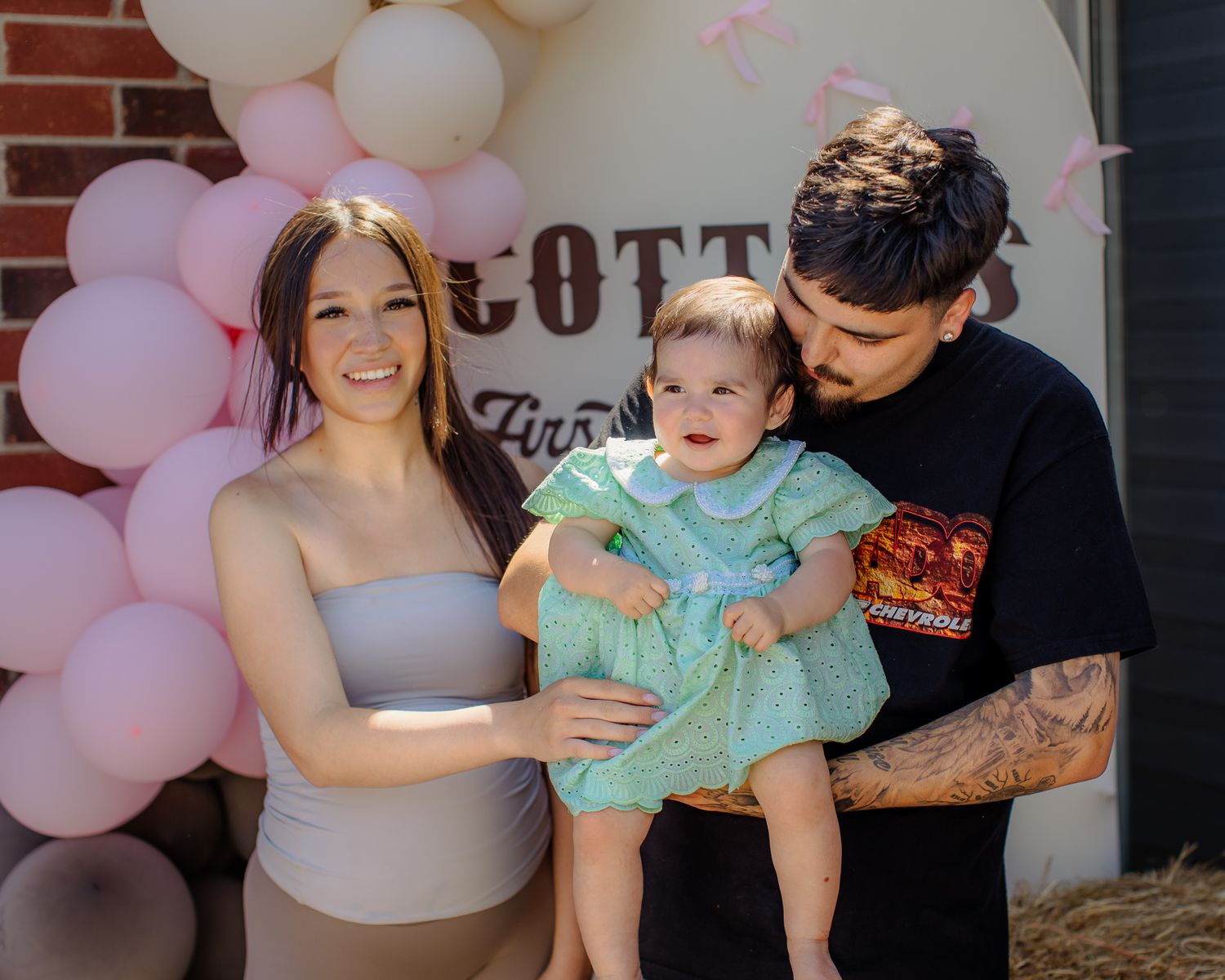 Family of three posing together at Scottie's first birthday rodeo themed party with balloon arch.