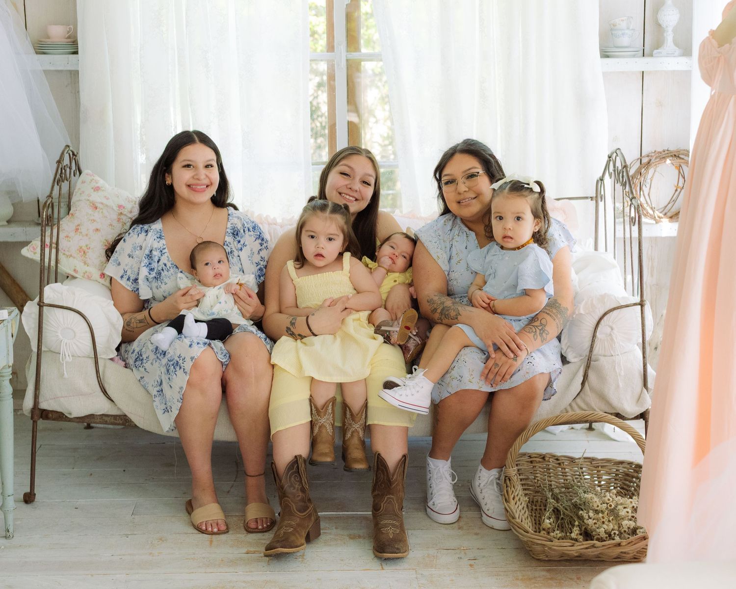 Three women sitting on a vintage sofa holding three babies in matching outfits in a bright, airy room.