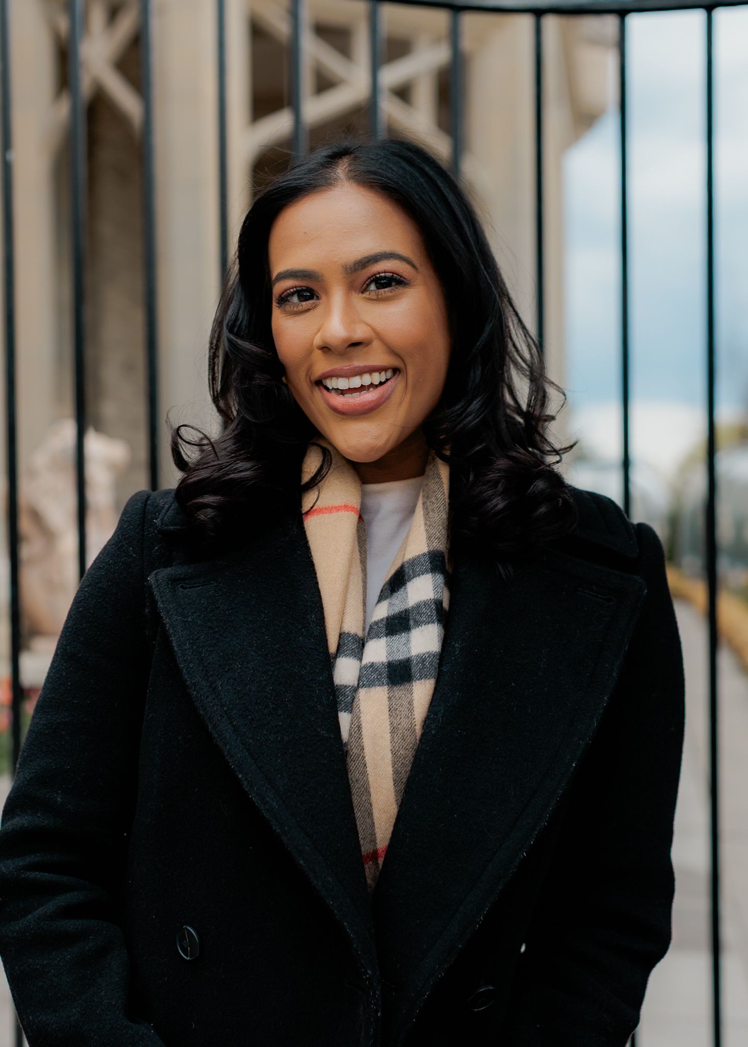 Smiling woman in black coat and Burberry scarf standing outdoors in front of iron gates.