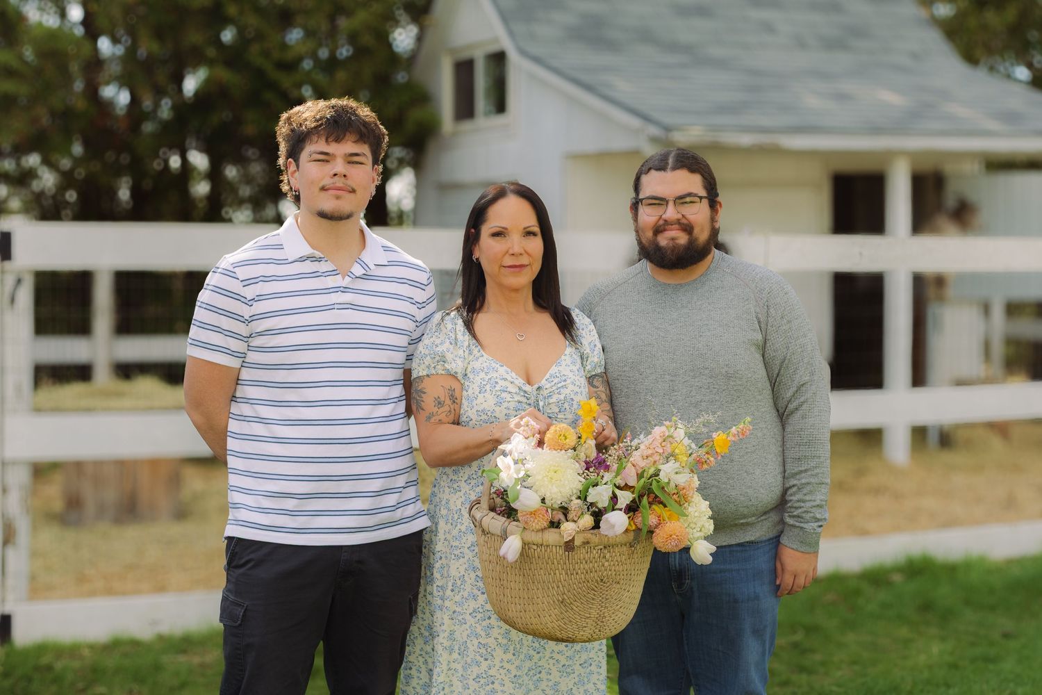 Three people posing outdoors near a white farmhouse, woman holding a basket of fresh wildflowers.