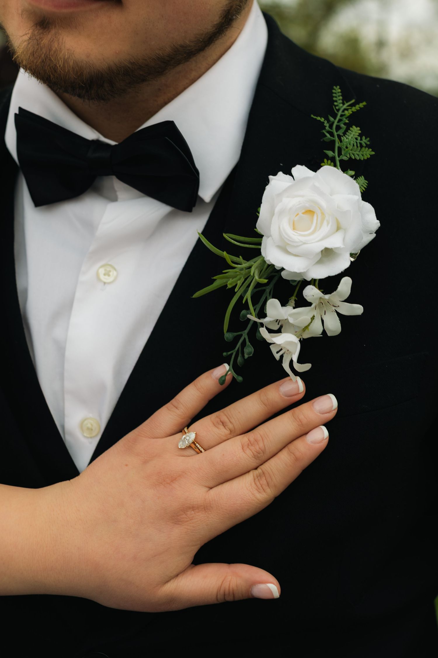 Bride places hand on grooms black tuxedo lapel featuring white rose boutonniere, showing engagement ring.