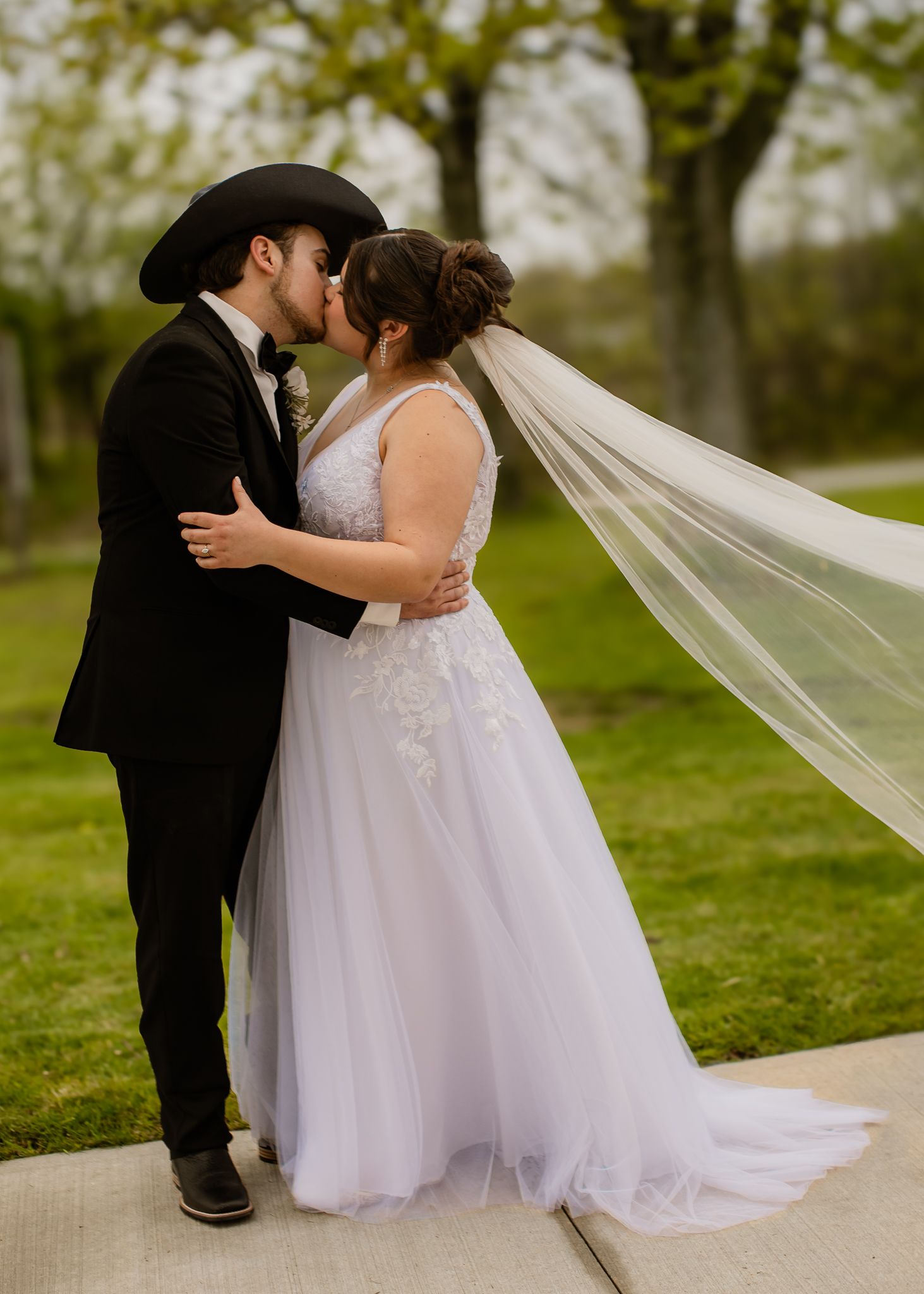 Bride and groom in cowboy hat share a kiss outdoors with long veil blowing in breeze.