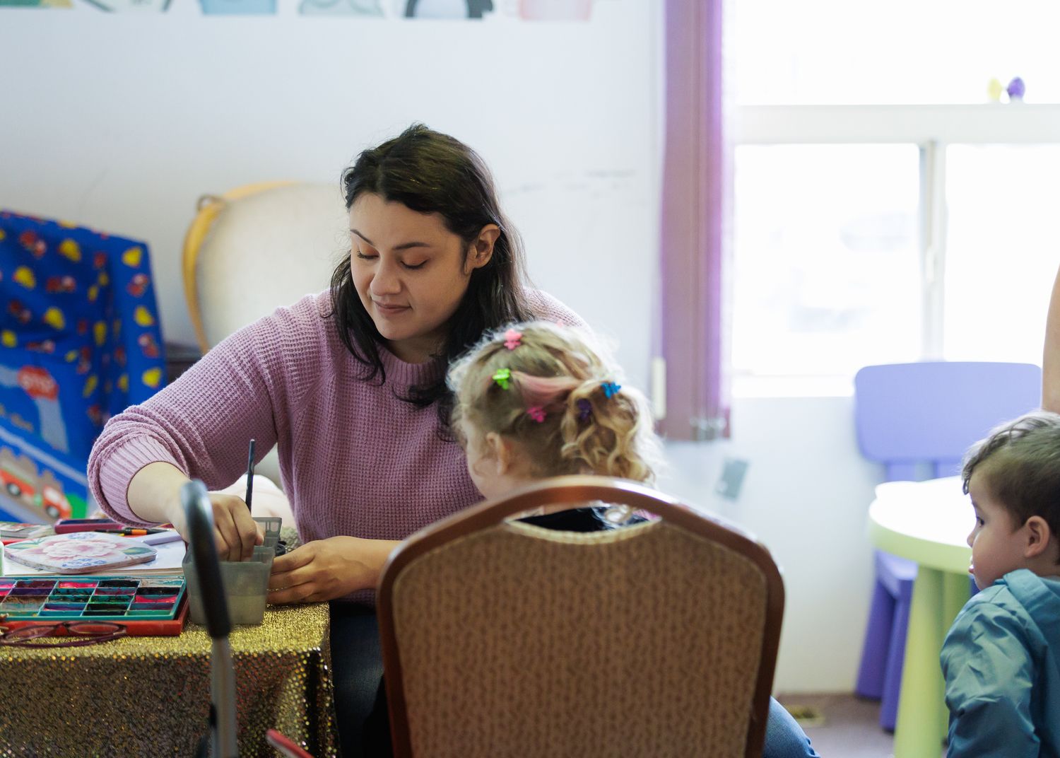 A woman helps a young child with an art activity at a table in a colorful classroom setting.