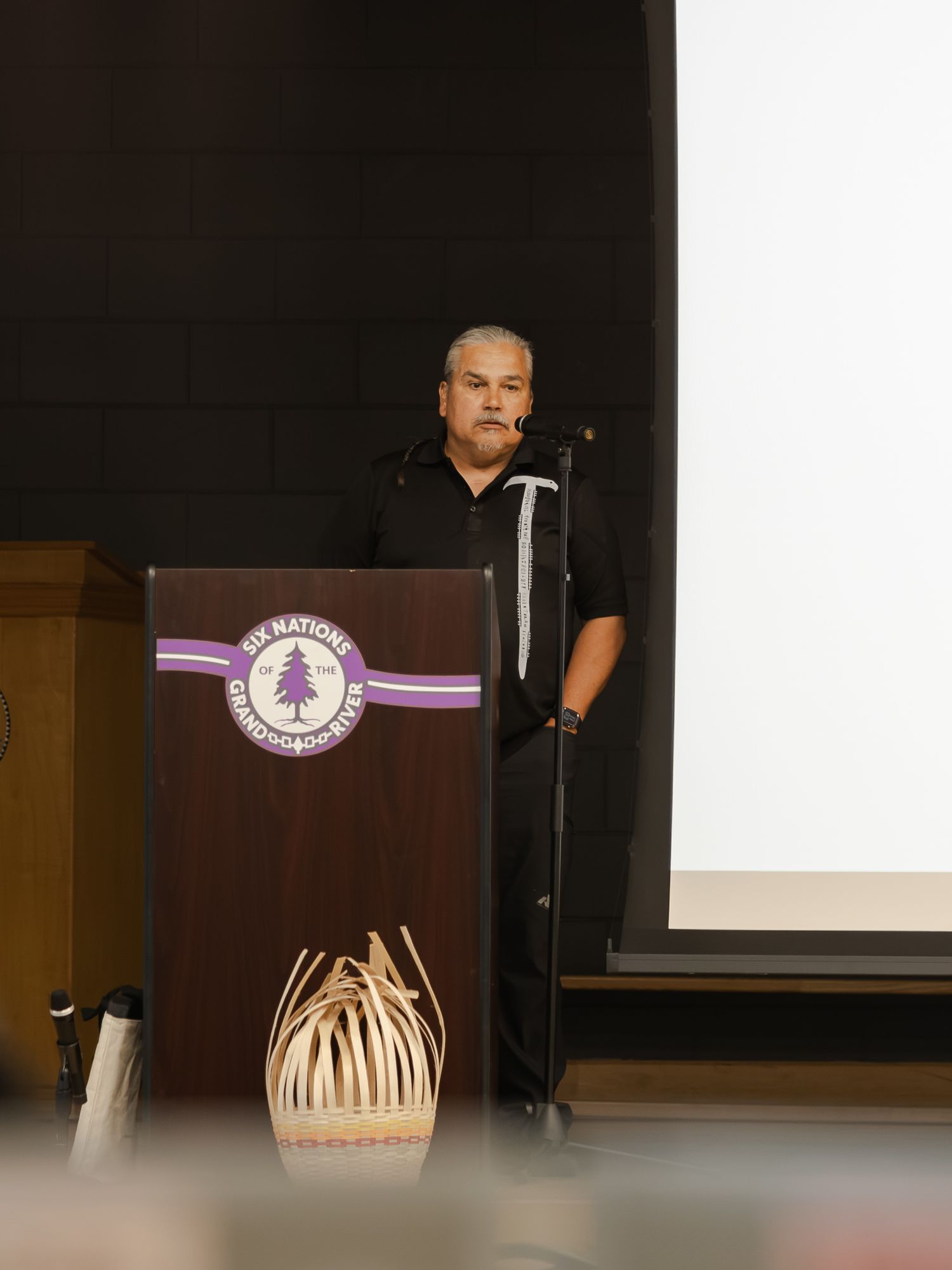 Man speaking at a Six Nations Ontario podium during a formal Indigenous community event or conference.