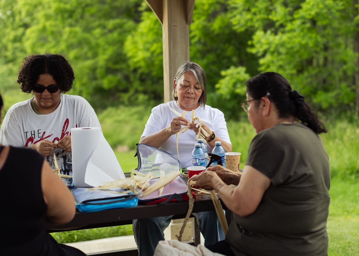 Three women sit at outdoor picnic table doing traditional Indigenous craftwork in green park.