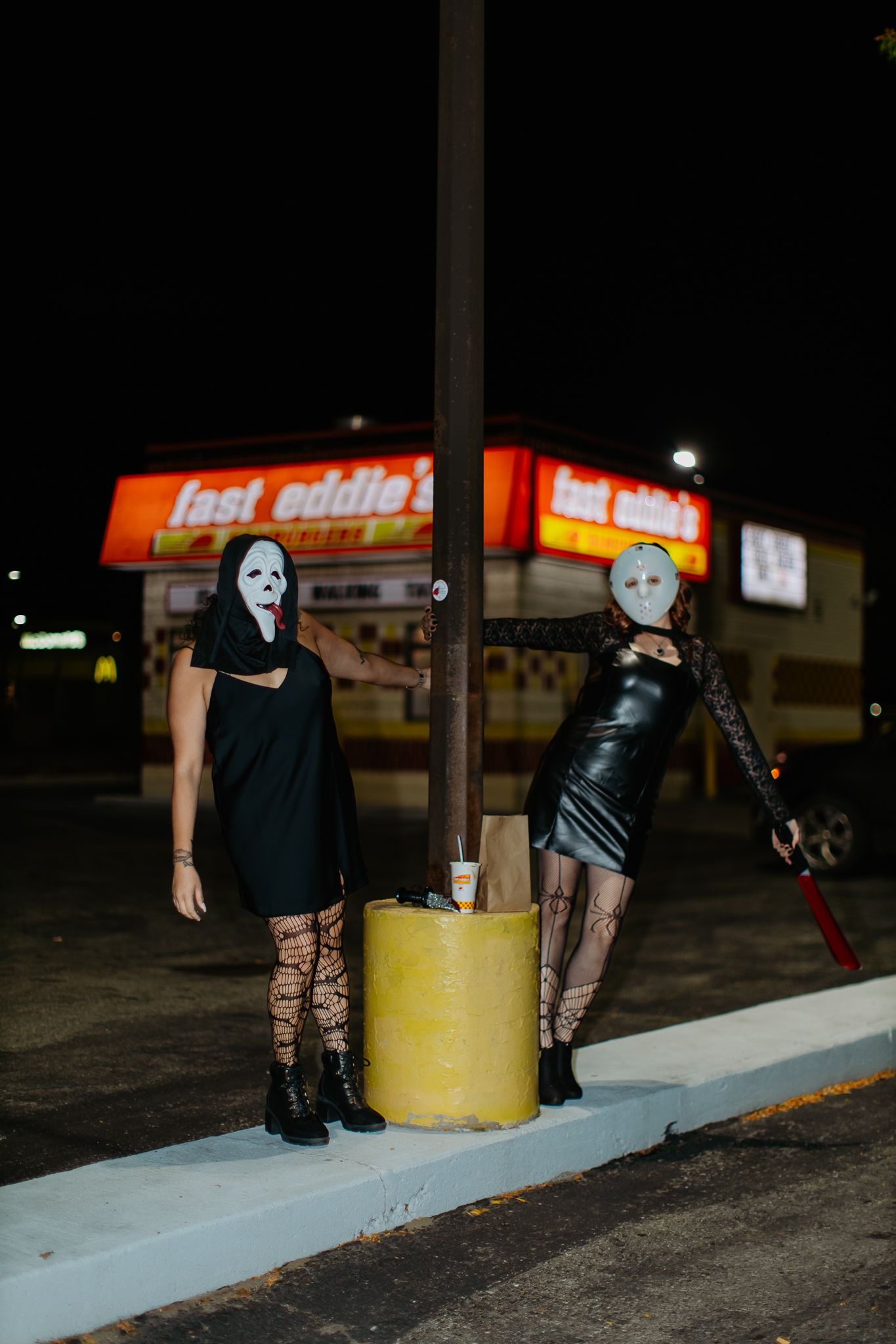 Dark nighttime photo showing figures posing near a yellow bollard and curb outside Fast Eddie's restaurant.