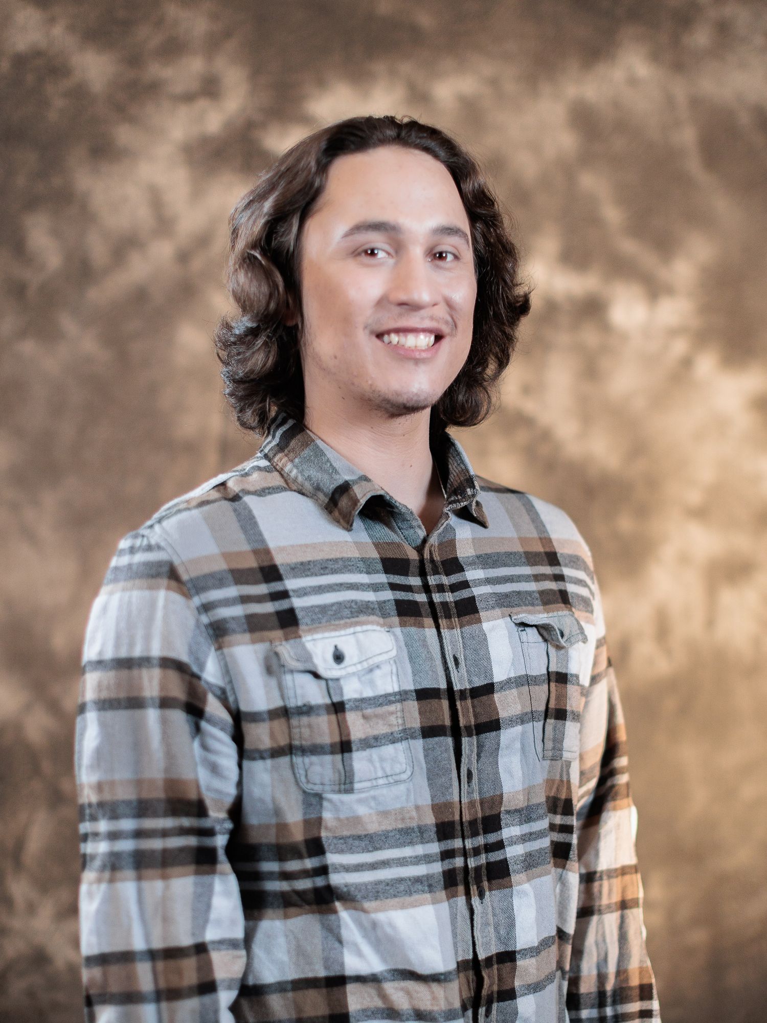 Young man with shoulder-length hair smiling, wearing a plaid flannel shirt against a brown studio backdrop.