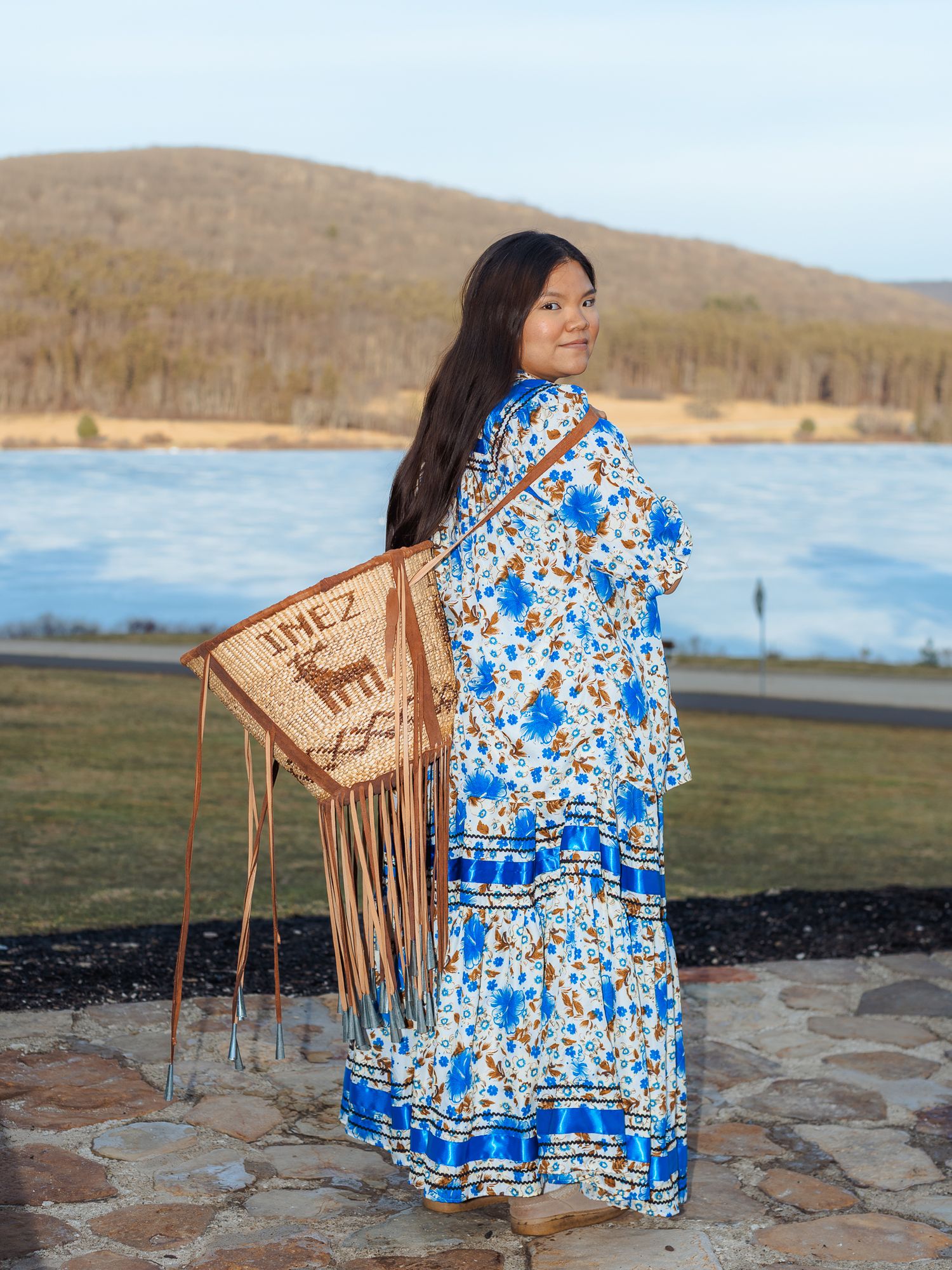 Young woman in blue floral dress holding a fringed leather bag with deer motif, standing by a scenic lake and mountain backdrop.