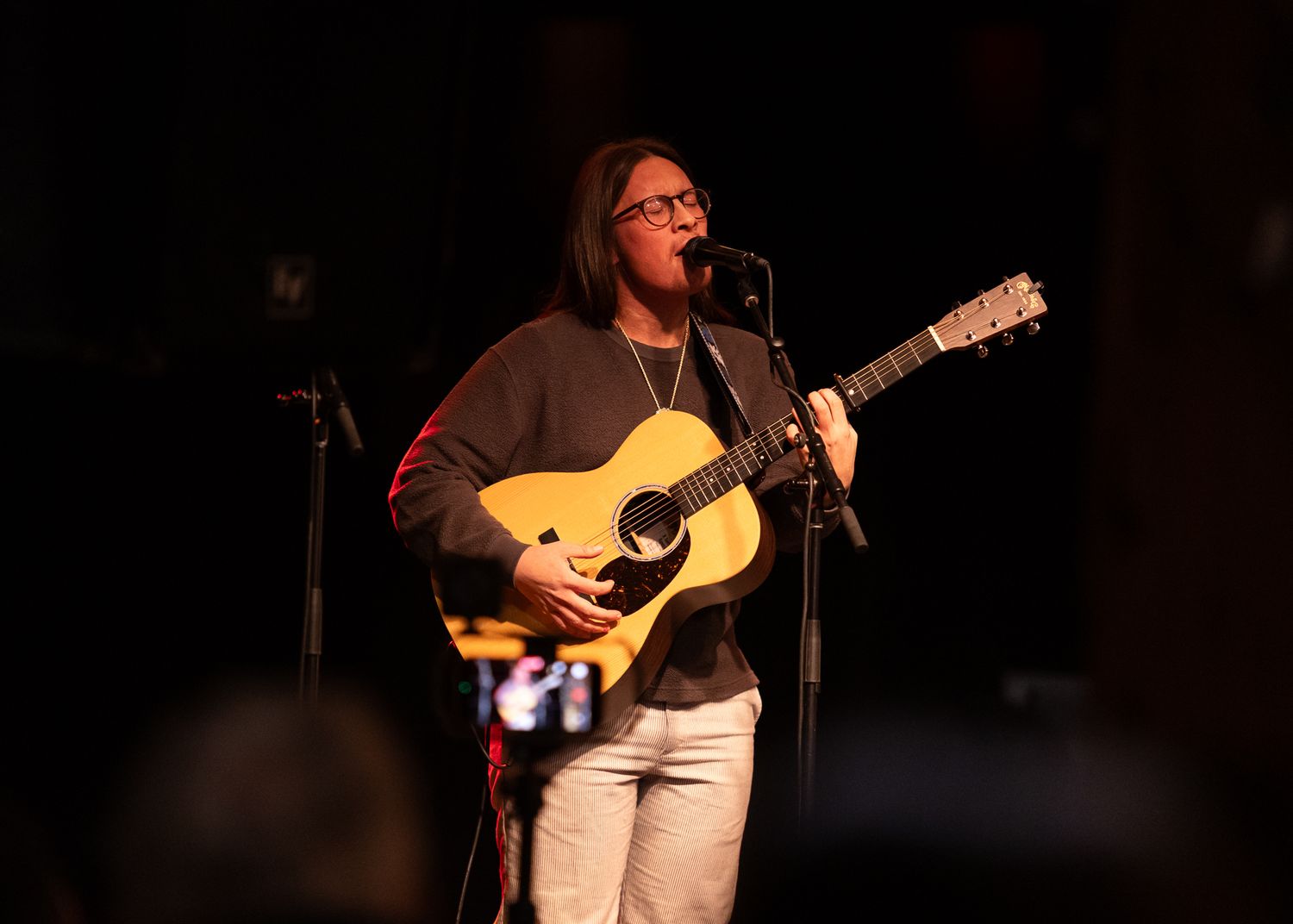 Young woman playing acoustic guitar and singing into a microphone on a dimly lit stage.