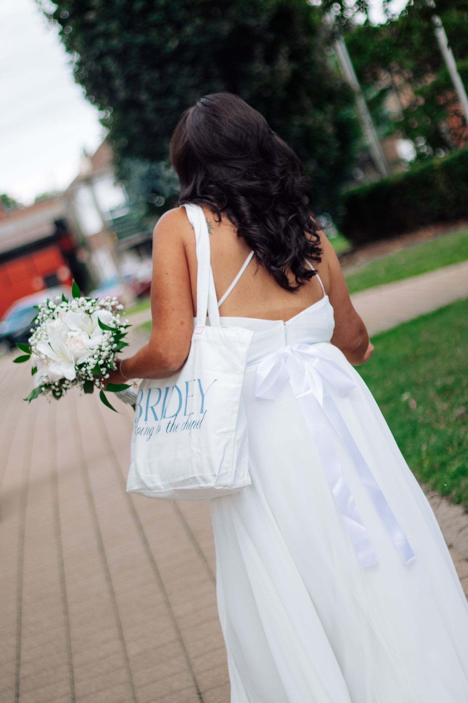 Bride in backless white wedding gown with bow carries bouquet and tote bag while walking along an outdoor pathway.