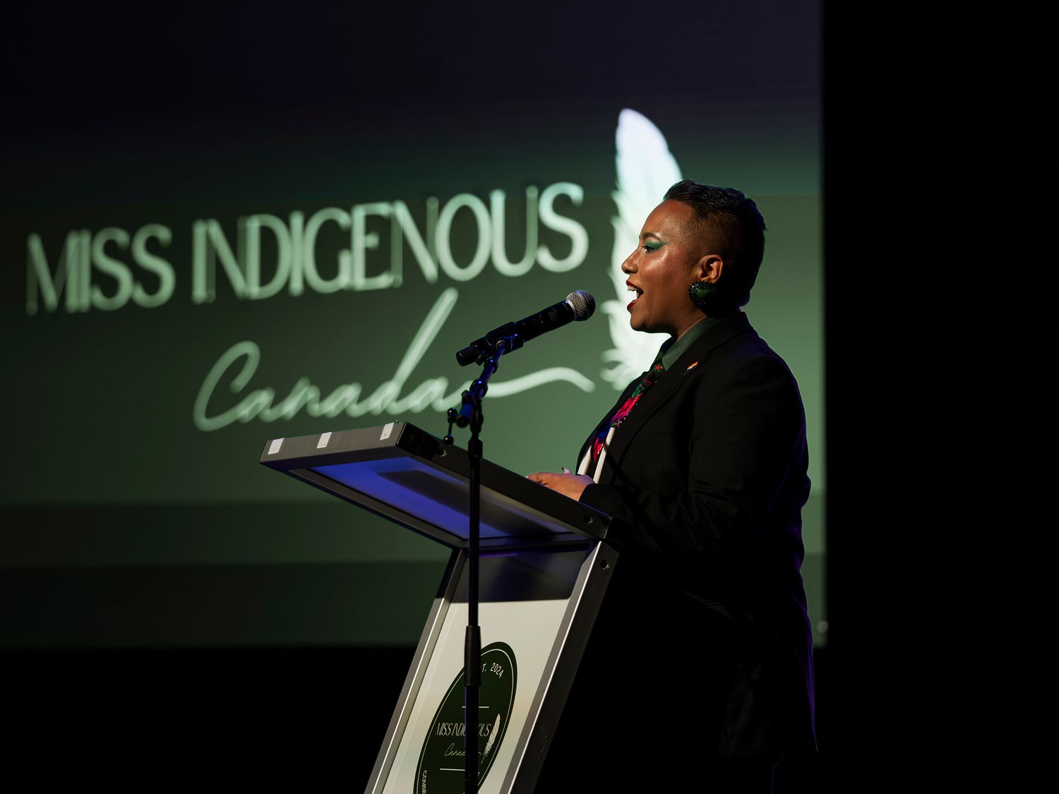 Speaker at podium during Miss Indigenous Canada pageant event with logo projected on screen.