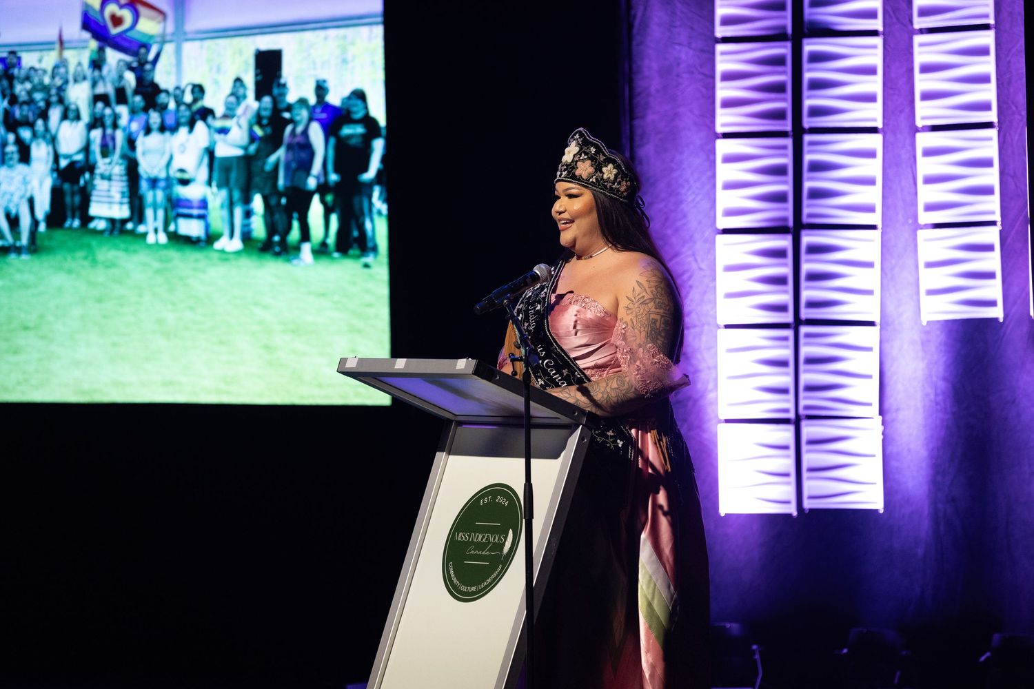 A woman in an elegant gown and crown speaks at a podium during a formal awards ceremony with purple lighting.