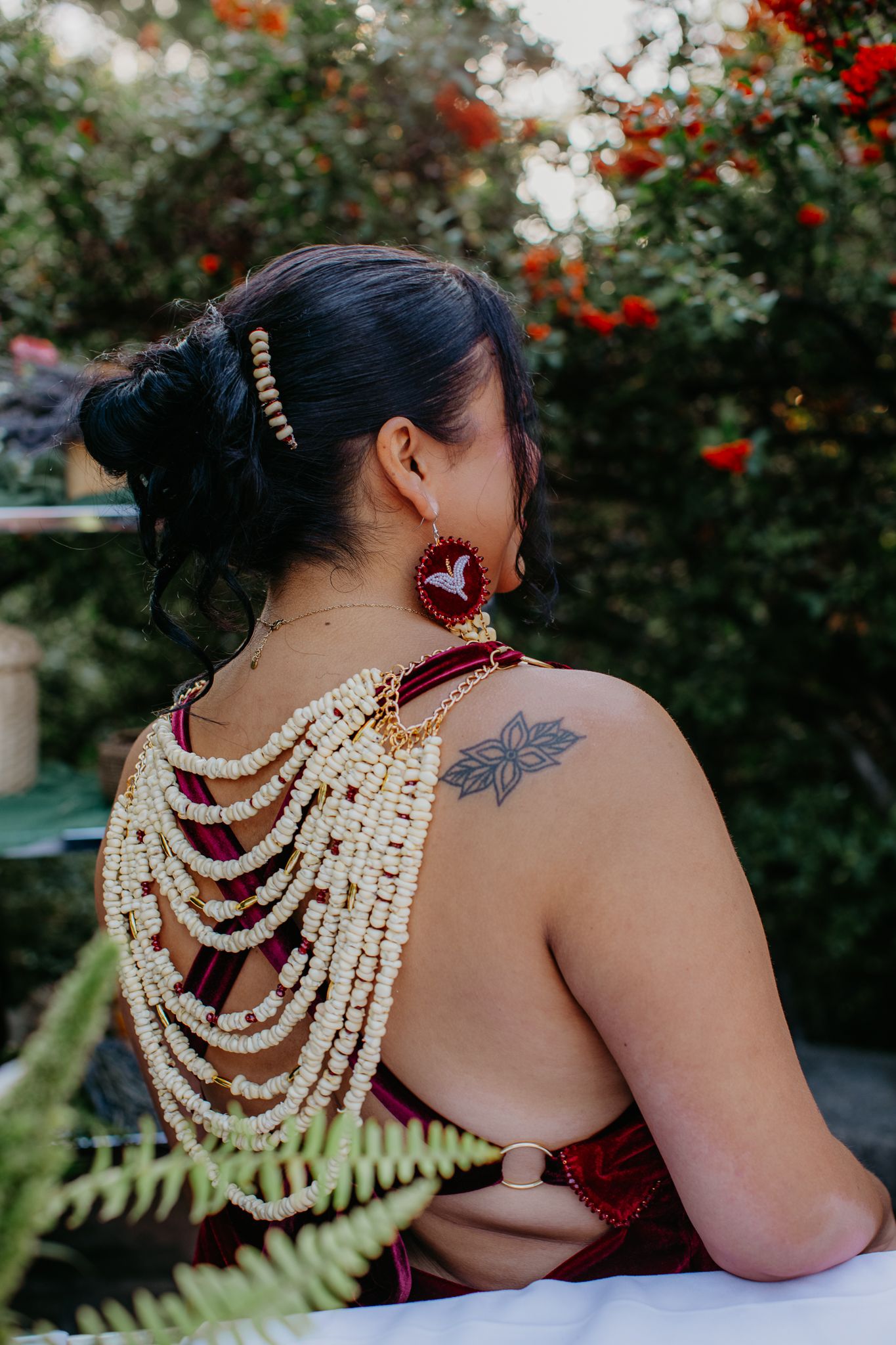 Woman with black updo hair, floral tattoo on shoulder, wearing beaded back necklace and red earrings outdoors.