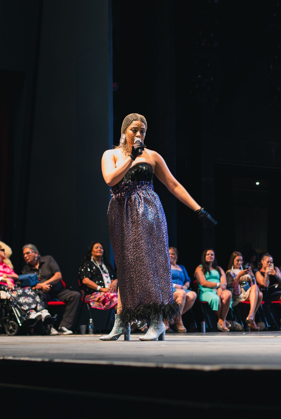 Woman in sequined gown performing on stage with microphone before seated audience at live event.