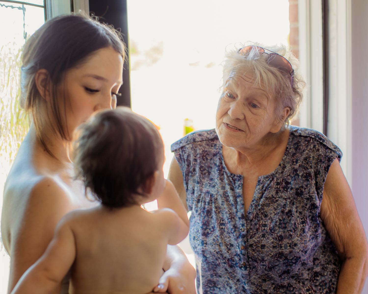 Young mother holds baby while elderly grandmother looks on warmly near a bright window.