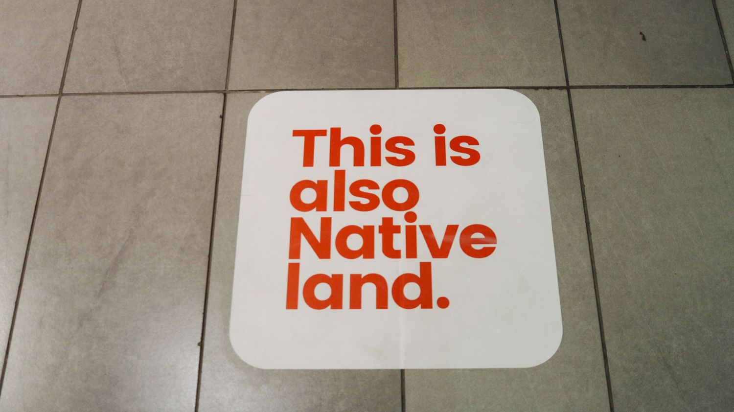 White floor sticker with bold orange text reading This is also Native land on tiled floor.