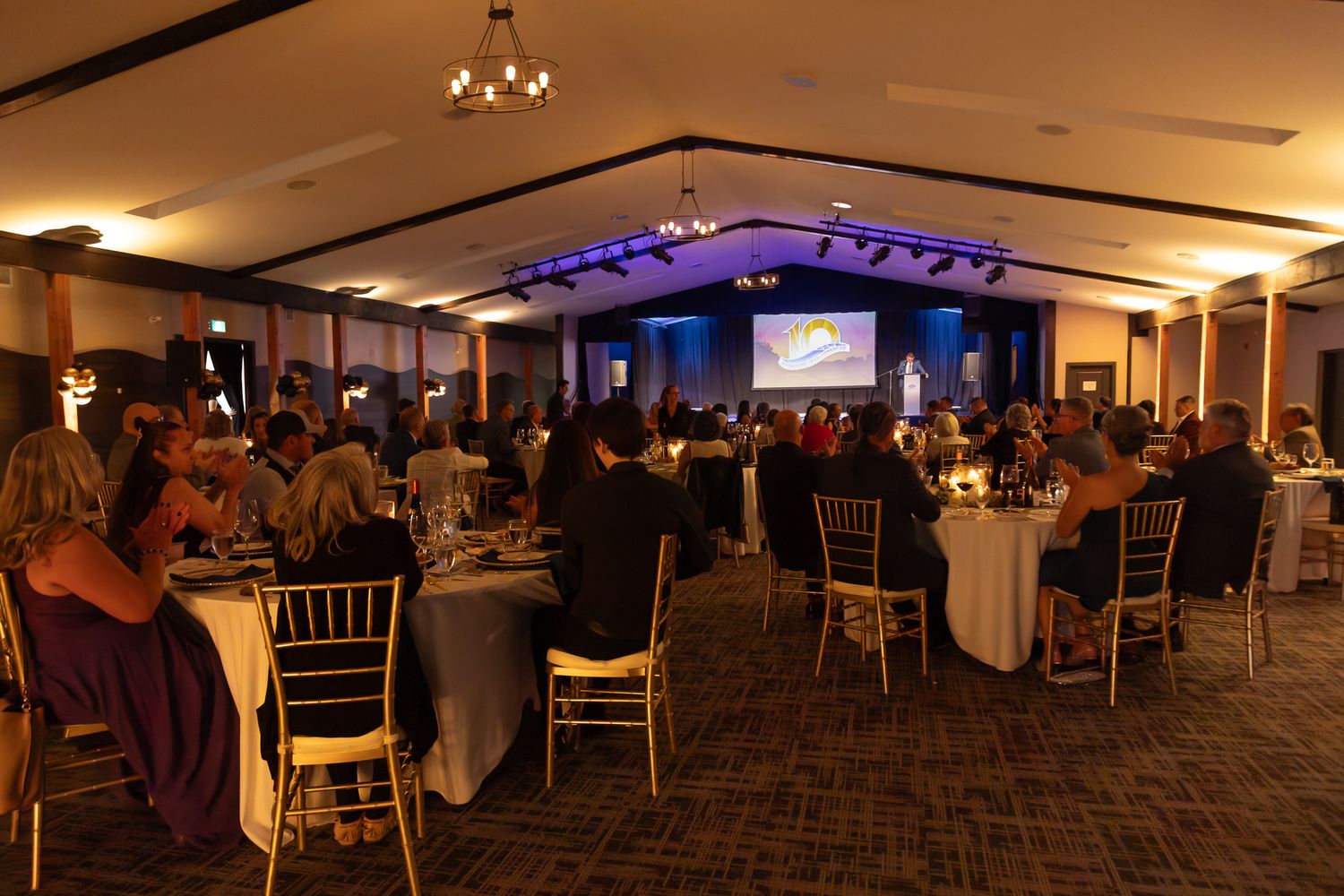 Elegant banquet hall with guests dining at round tables, gold chairs, and a glowing blue stage backdrop.