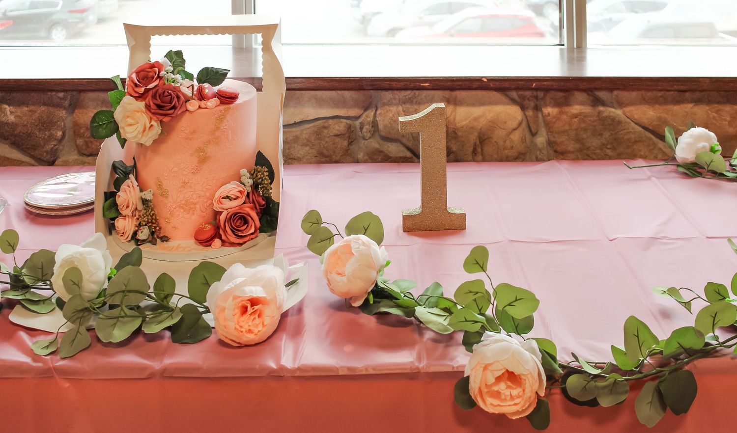 Elegant first birthday cake with pink floral decorations and greenery garland on a coral tablecloth with number one sign.