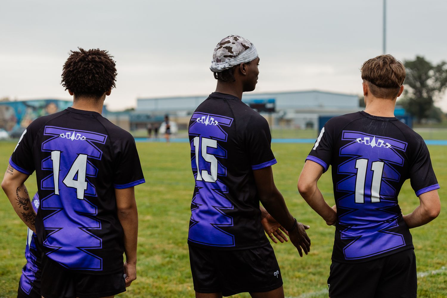 Three soccer players in purple and black jerseys numbered 14, 15, and 11 stand on a green field.