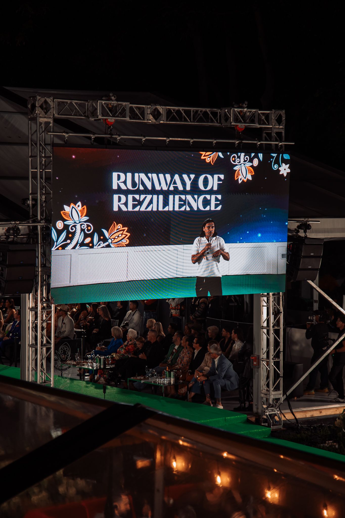 Large LED screen displaying Runway of Rezilience event with speaker on stage before a seated crowd at night.