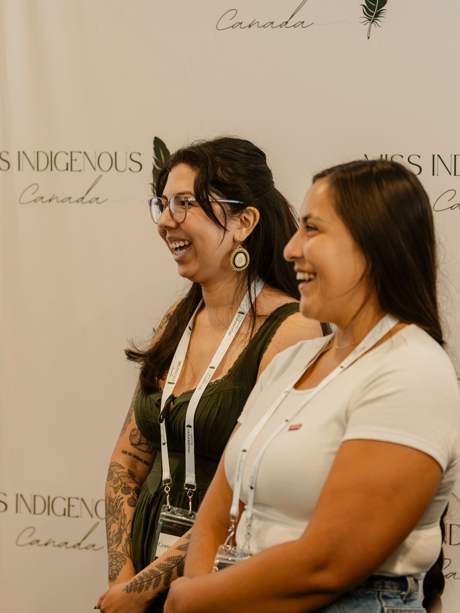 Two smiling women pose at a Miss Indigenous Canada event backdrop, one wearing glasses and a green top.