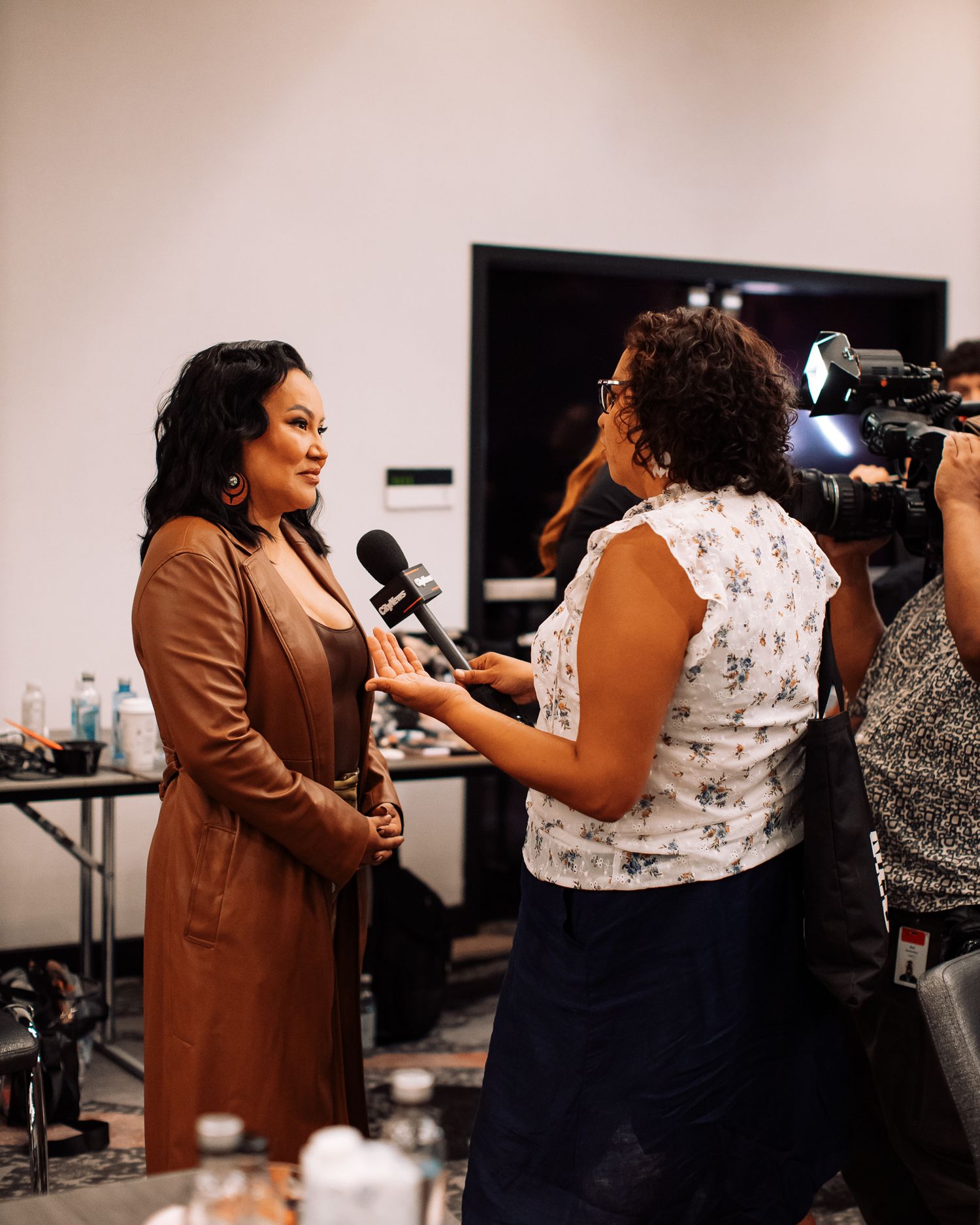 Woman in brown leather jacket being interviewed on camera at an indoor media event.