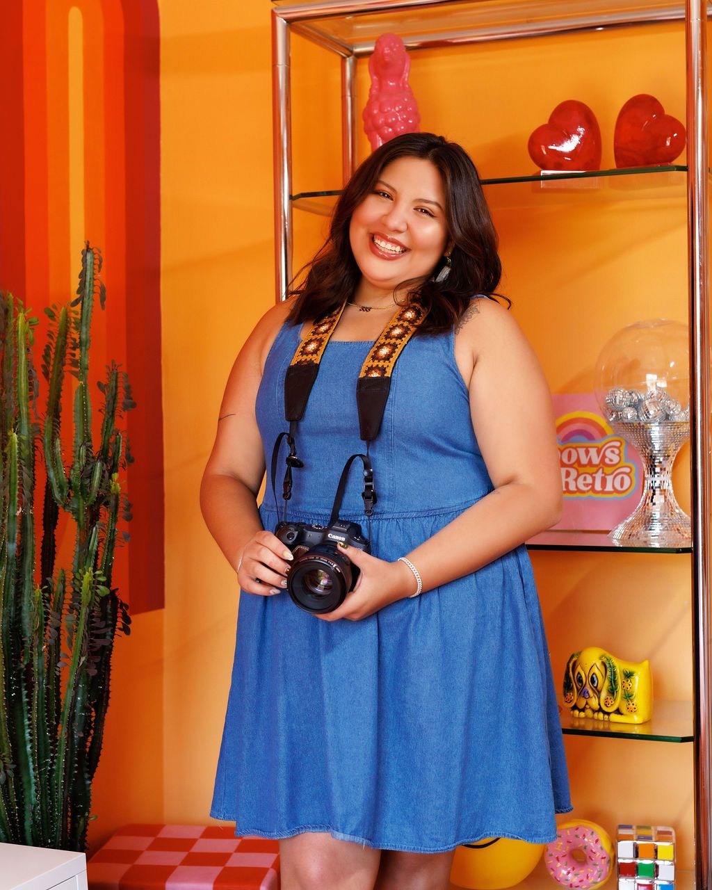 Woman in blue dress holding camera smiling in colorful retro orange room with shelves and cactus.