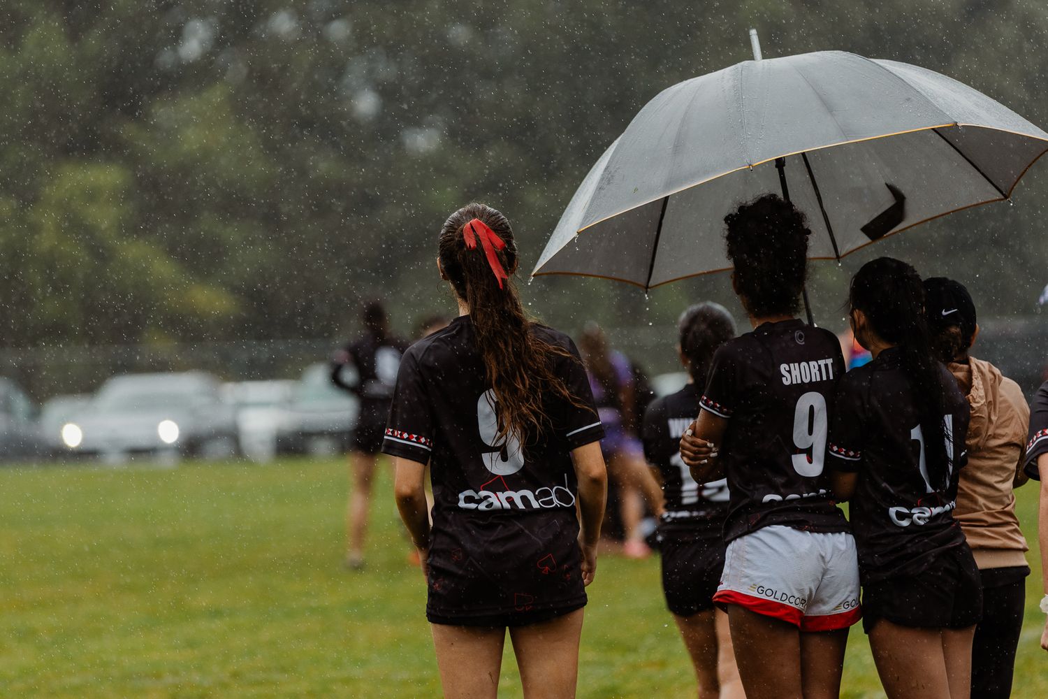 Female rugby players in black jerseys walk in the rain sharing an umbrella during a wet outdoor match.