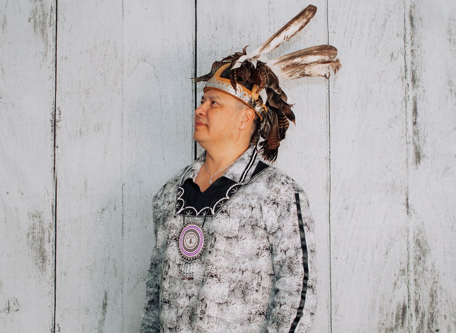 A person wearing traditional Native American regalia with feathers poses against a white wooden wall.
