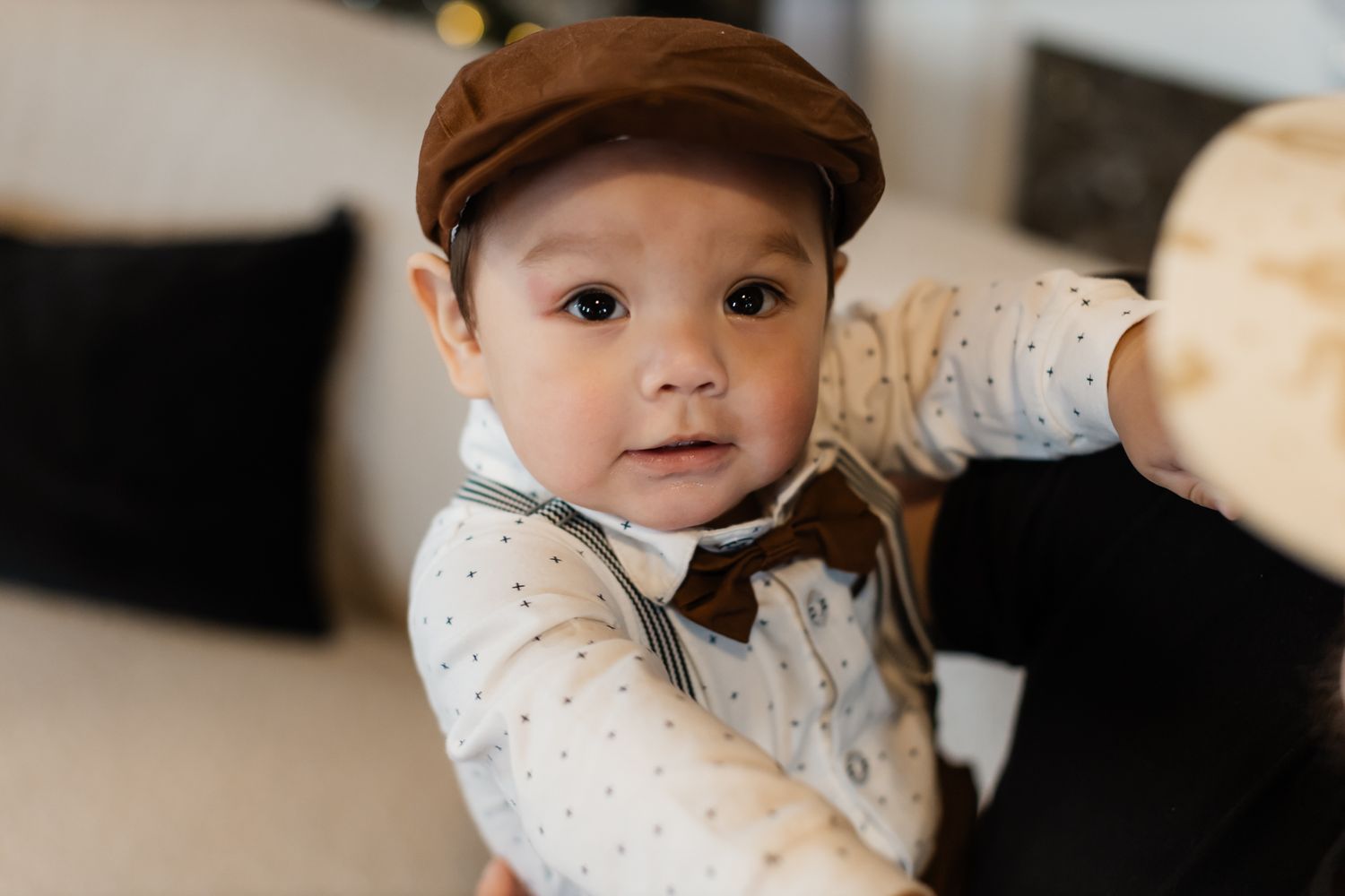 Young toddler wearing a newsboy cap and bowtie looks at camera with curious expression.