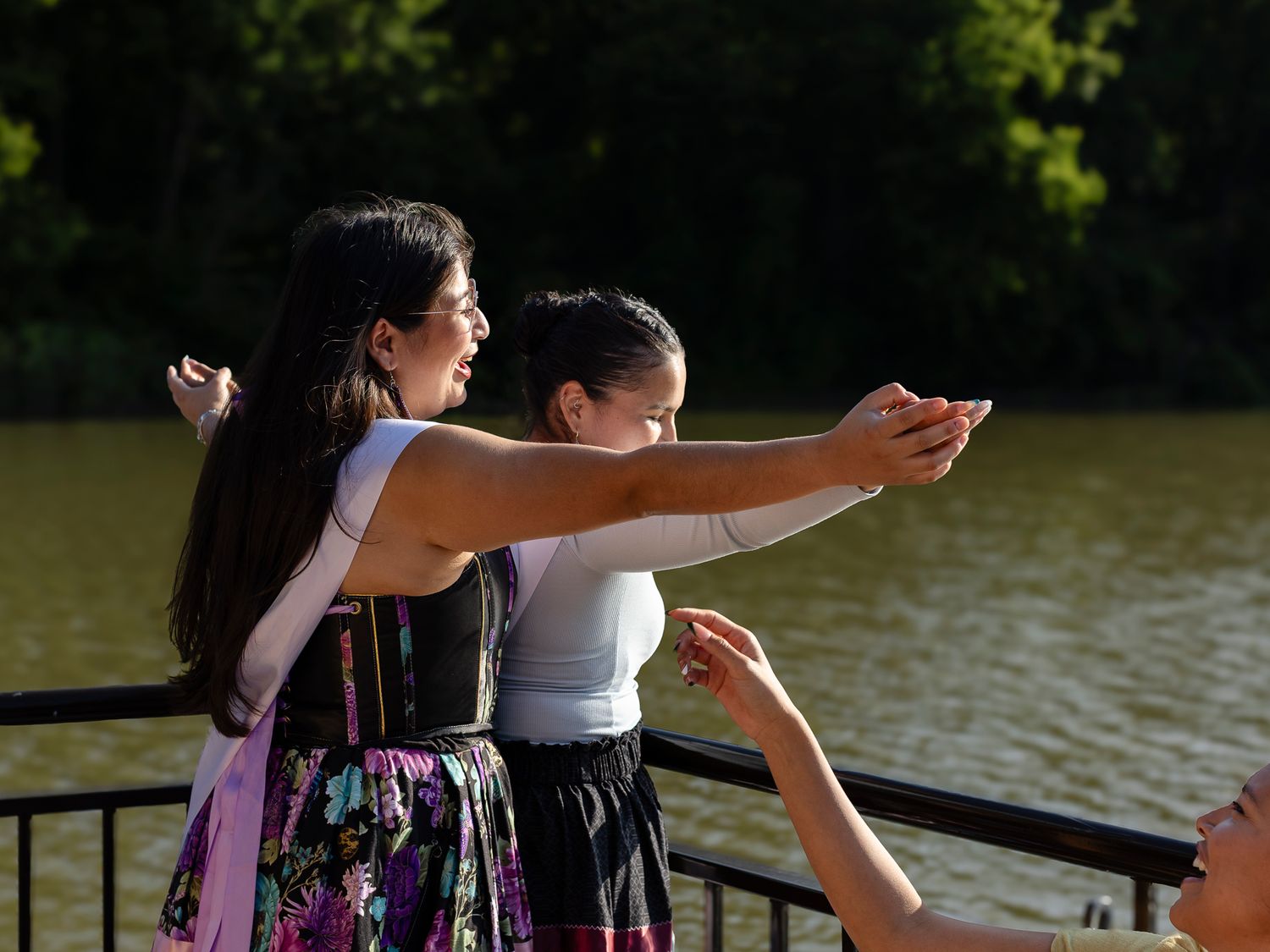 Two women lean over a railing taking a selfie by a sunlit lake surrounded by lush green trees.