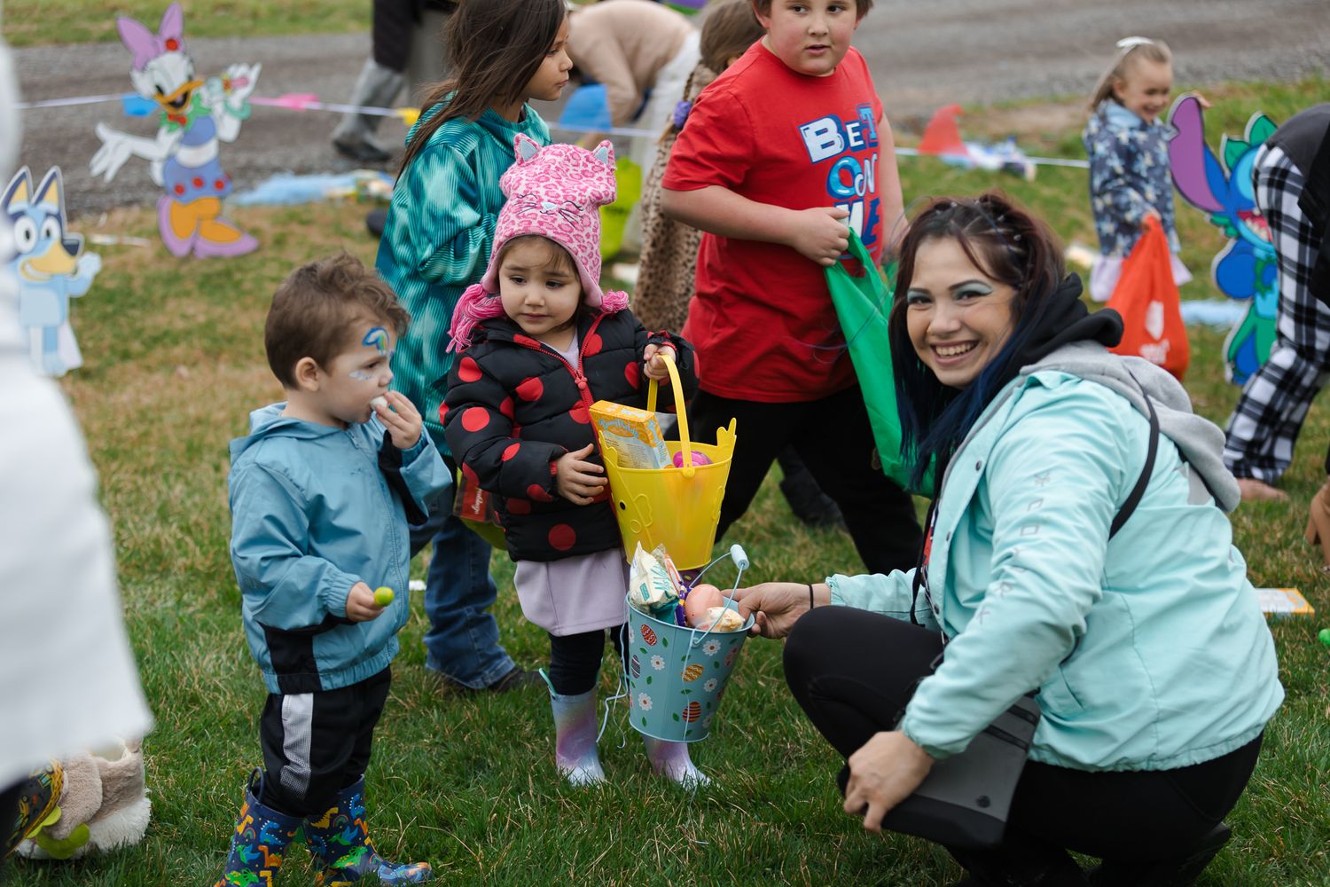 Woman crouches with two toddlers holding Easter baskets at an outdoor egg hunt event with other children nearby.