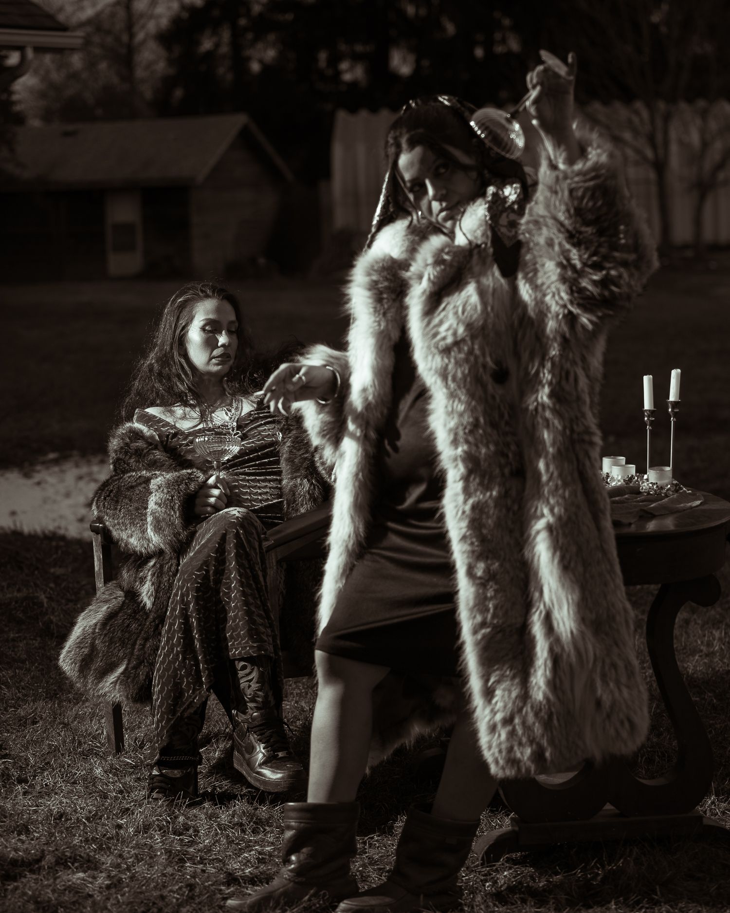 Two women in fur coats gathered around outdoor table with candles in black and white photo.