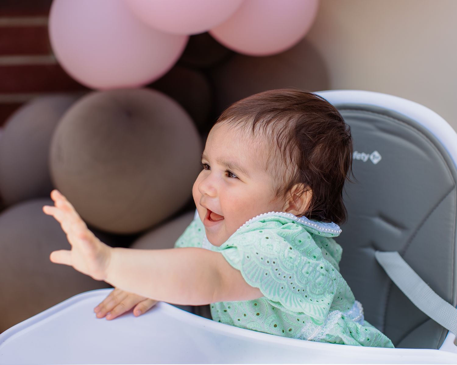 Happy baby in a high chair wearing a mint green outfit reaching out at a birthday party.